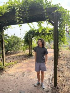 A young person stands wearing athletic clothing in a garden under a pergola draped in vines. 